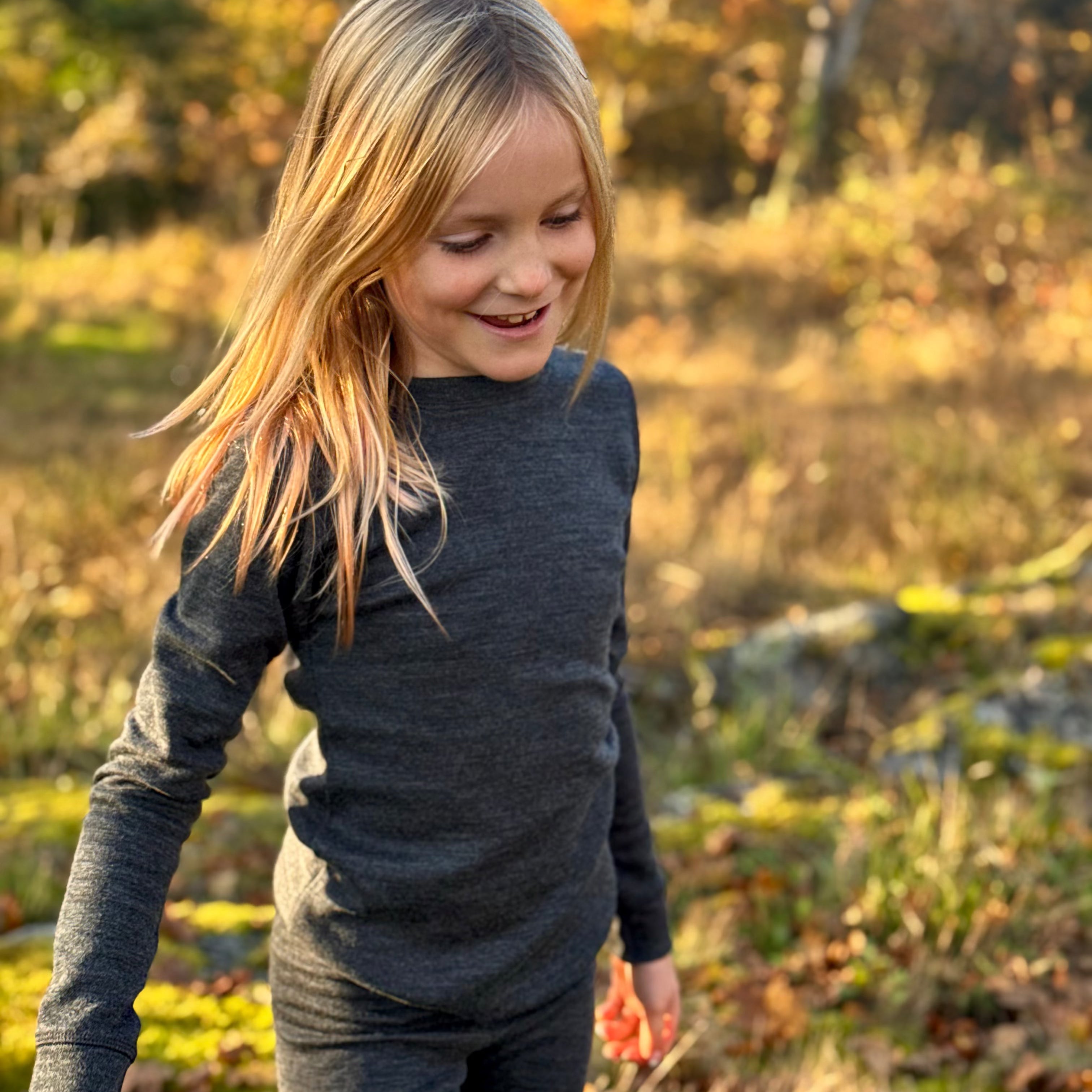 Child in a dark merino wool long-sleeve shirt and pants standing in a field with trees in the background.
