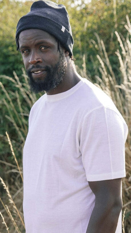 A man wearing a gray basic T-shirt and brown pants standing near a rocky beach with driftwood.